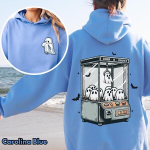 Pode incluir: Um moletom azul claro com um gr&aacute;fico de uma m&aacute;quina de garras fantasma com o texto "Grab-A-Ghost". O moletom tem um pequeno gr&aacute;fico de fantasma na frente.