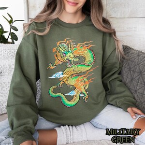 K&ouml;nnte beinhalten: Ein gr&uuml;nes Sweatshirt mit einem grafischen Aufdruck eines gelben und gr&uuml;nen Drachen mit orangefarbenen Flammen. Der Drache ist zu einem Kreis zusammengerollt, mit dem Kopf zum Betrachter gewandt.