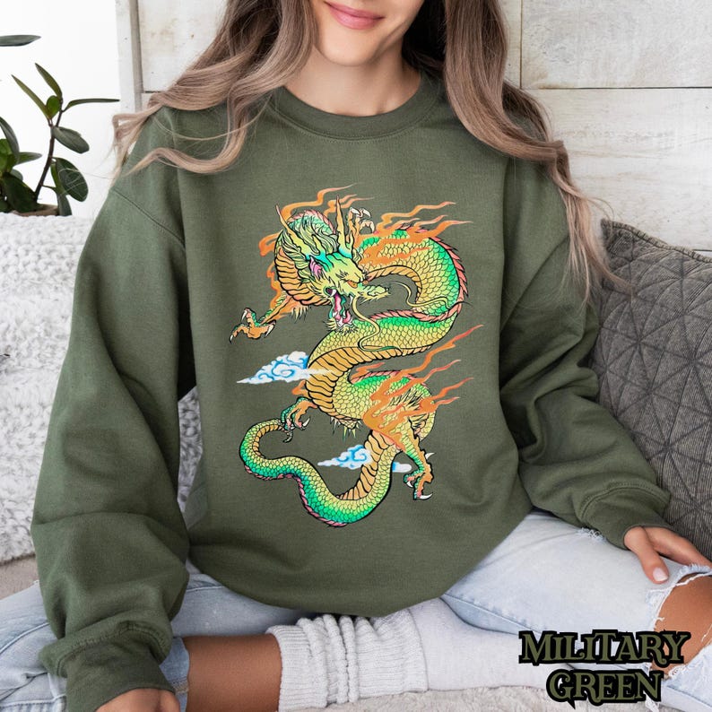 K&ouml;nnte beinhalten: Ein gr&uuml;nes Sweatshirt mit einem grafischen Aufdruck eines gelben und gr&uuml;nen Drachen mit orangefarbenen Flammen. Der Drache ist zu einem Kreis zusammengerollt, mit dem Kopf zum Betrachter gewandt.