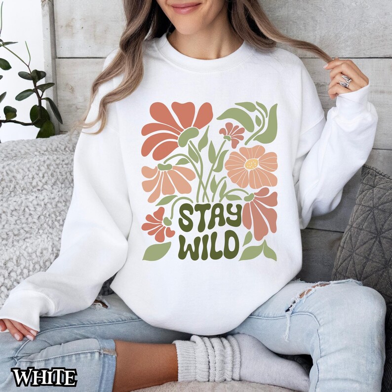 May include: White sweatshirt featuring a floral design in orange, peach, and green. The words "STAY WILD" are printed in green. The sweatshirt has a crew neck.