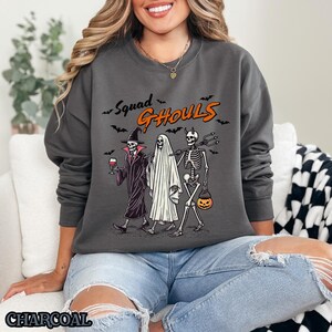 Pu&ograve; includere: Una felpa grigia antracite con una stampa grafica di tre scheletri vestiti in costume. Il testo "Squad Ghouls" &egrave; stampato sopra gli scheletri.