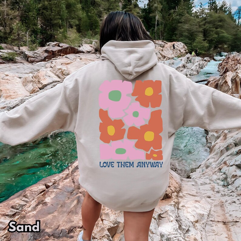 May include: A sand-colored hooded sweatshirt featuring a large graphic on the back. The design consists of stylized pink and orange flowers with yellow and green centers. Below the floral graphic, the text reads LOVE THEM ANYWAY in blue.