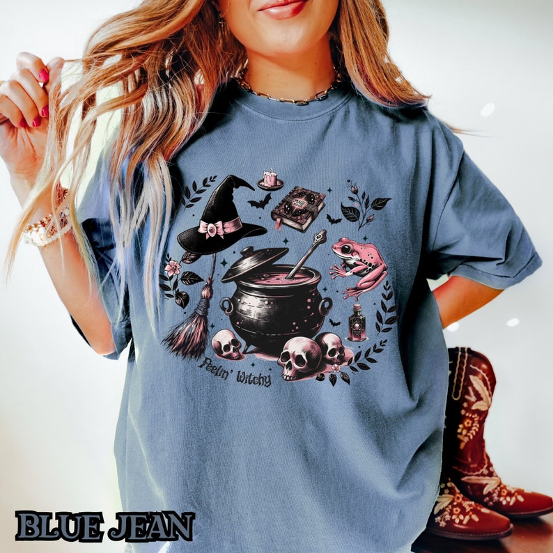 May include: A blue t-shirt with a graphic print of a witch's cauldron, a frog, a broom, a book, and a skull. The text "Feelin' Witchy" is printed below the graphic.