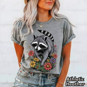 May include: A gray t-shirt with a graphic of a raccoon surrounded by colorful flowers. The raccoon is gray and black with a white face and black eyes. The flowers are red, yellow, purple, and pink.