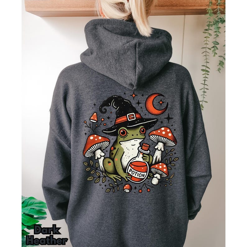 May include: Dark heather grey hoodie featuring a graphic of a frog wearing a witch's hat and holding a potion bottle. The frog is surrounded by mushrooms and stars.