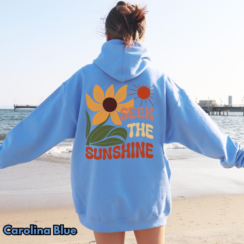 K&ouml;nnte beinhalten: Ein hellblauer Hoodie mit einer Sonnenblume und dem Text "SEEK THE SUNSHINE" in einer Retro-Schrift. Der Hoodie hat eine Kapuze mit Kordelzug und eine K&auml;ngurutasche.