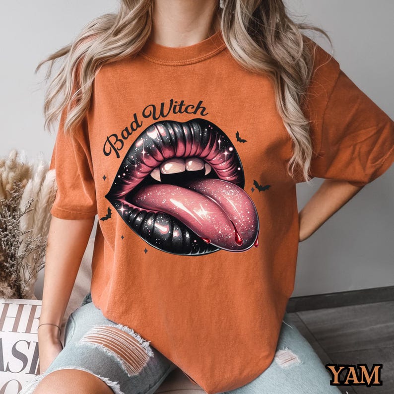 Peut inclure: T-shirt orange avec un graphique noir et rose d'une bouche avec des crocs et une langue qui sort. Le texte "Bad Witch" est au-dessus du graphique.