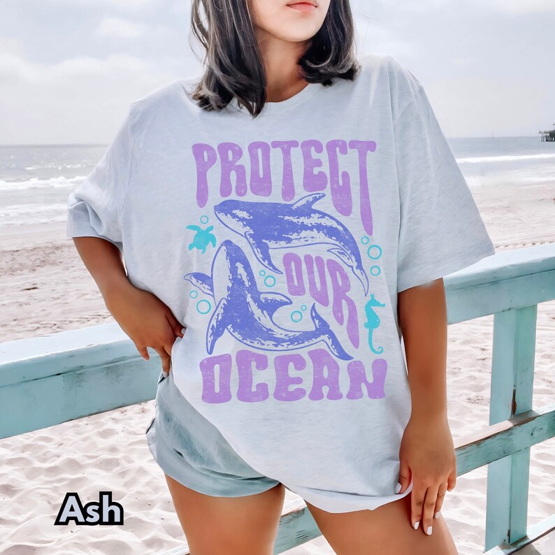 Pu&ograve; includere: Una maglietta blu chiaro con una stampa grafica di due balene e il testo "PROTECT OUR OCEAN" in un font viola retr&ograve;. La grafica include anche una tartaruga marina e un cavalluccio marino.