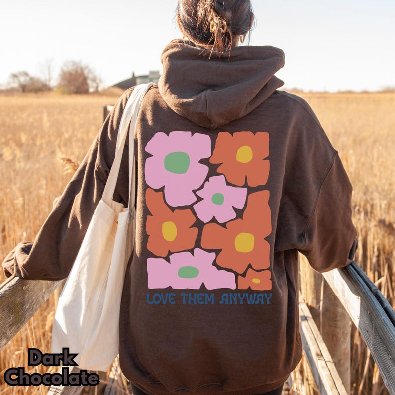 May include: A dark chocolate brown hooded sweatshirt featuring a large graphic on the back with stylized pink and orange flowers with yellow and green centers. The text "LOVE THEM ANYWAY" is printed in blue below the floral design.