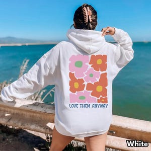 May include: A white hooded sweatshirt featuring a large rectangular graphic on the back with stylized pink and orange flowers with yellow and green centers. The text LOVE THEM ANYWAY is printed in blue below the floral design.