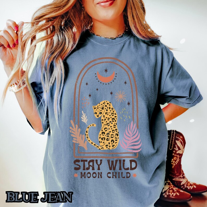 May include: A blue short-sleeve t-shirt featuring a graphic print of a leopard inside an arched frame with a crescent moon, stars, and foliage. The text "STAY WILD MOON CHILD" is printed in brown below the leopard. The shirt has a crew neckline.