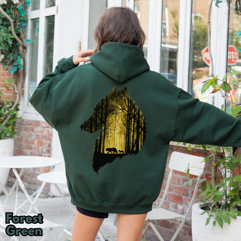 May include: A forest green hooded sweatshirt featuring a large graphic on the back. The graphic is a silhouette of a wolf head filled with a yellow-toned forest scene, depicting a wolf and a smaller pup walking among tall, dark tree trunks.
