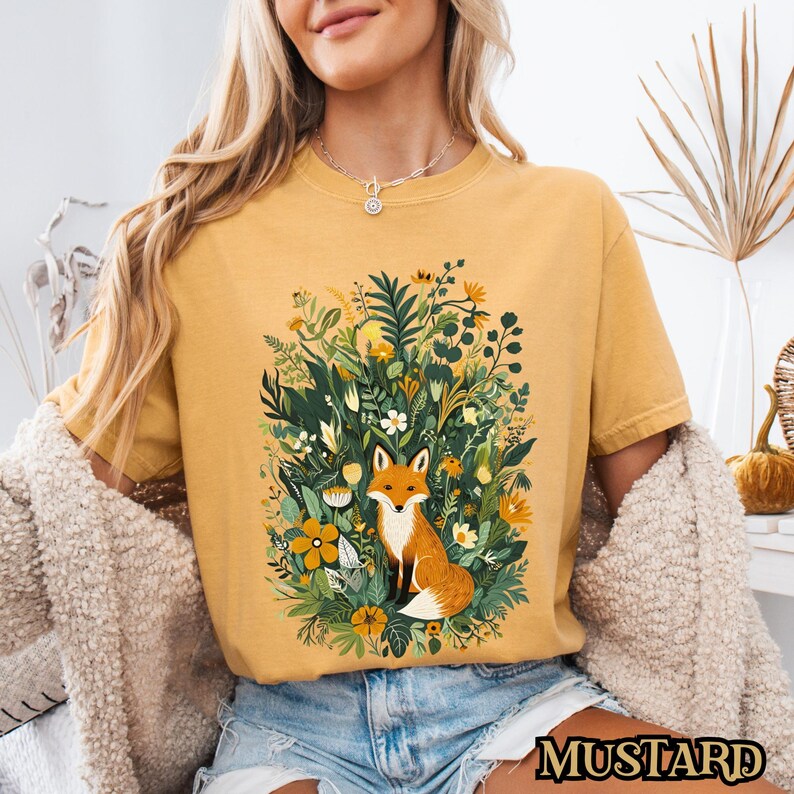 May include: A mustard yellow t-shirt with a graphic of a fox surrounded by flowers and greenery. The fox is orange and white with a bushy tail.