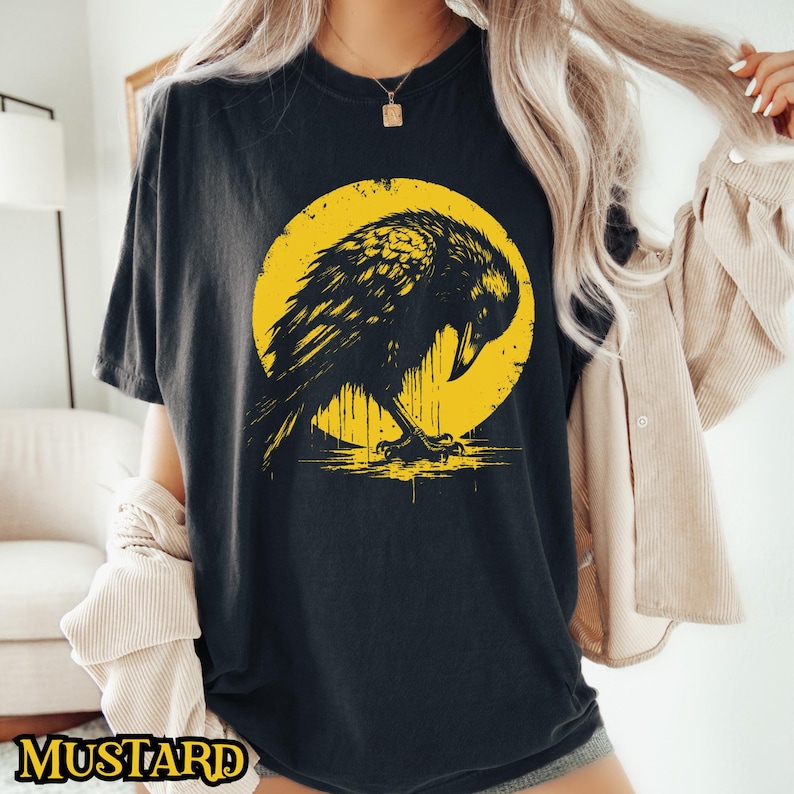 May include: Black t-shirt with a graphic of a black crow perched on a yellow moon. The crow is facing left and has its wings spread out.