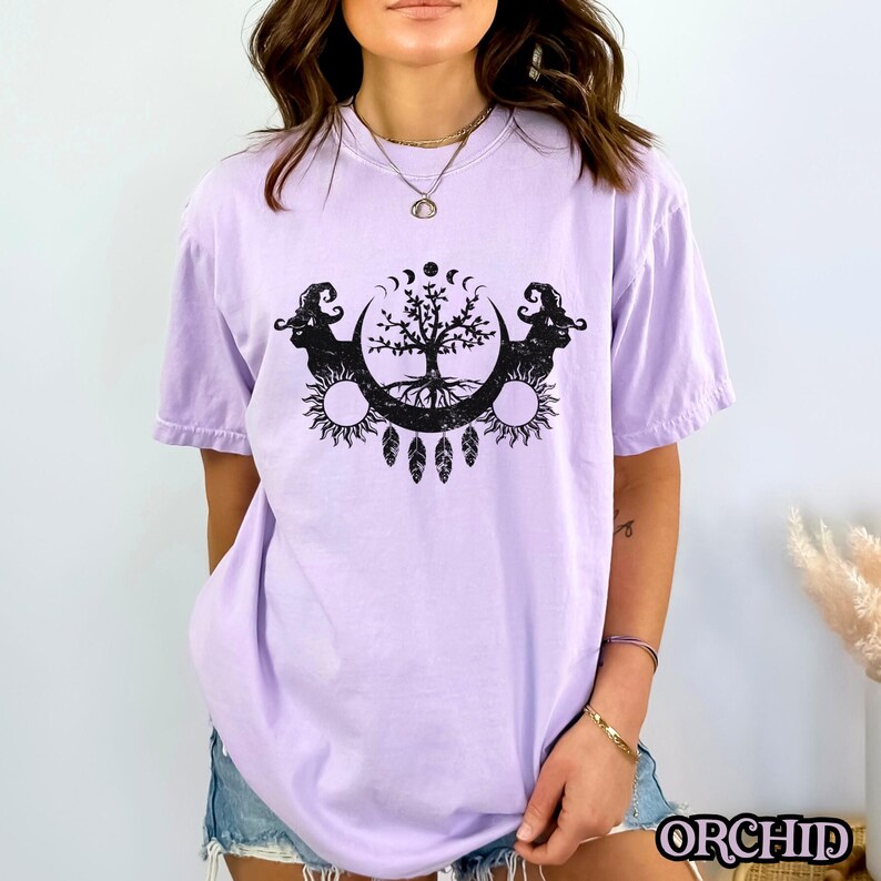 May include: A lavender t-shirt with a black and white graphic print of a tree with roots, surrounded by moons and suns. The graphic also includes two stylized animal heads.