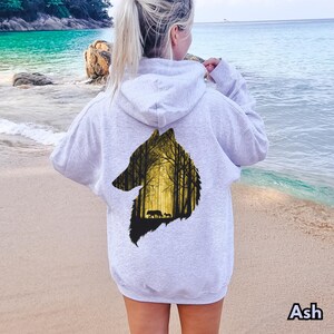 May include: A light gray hooded sweatshirt featuring a large graphic on the back. The graphic is shaped like a wolf head silhouette and contains a detailed illustration of a forest with tall trees in shades of yellow and black, including two wolves.