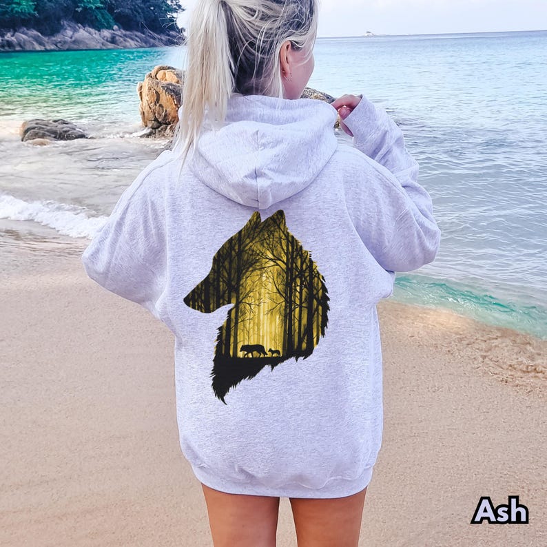 May include: A light gray hooded sweatshirt featuring a large graphic on the back. The graphic is shaped like a wolf head silhouette and contains a detailed illustration of a forest with tall trees in shades of yellow and black, including two wolves.