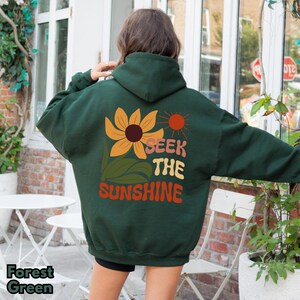 K&ouml;nnte beinhalten: Ein dunkelgr&uuml;ner Hoodie mit einer Sonnenblume, einer Sonne und dem Text "Seek the Sunshine" auf der R&uuml;ckseite.