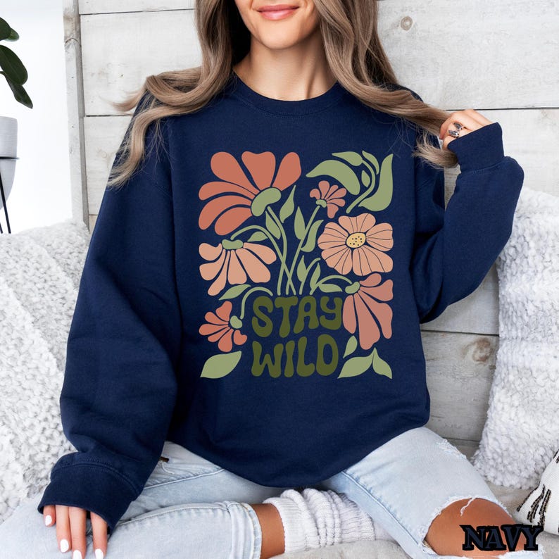May include: Navy blue sweatshirt with a floral design. The design includes peach and orange flowers, green leaves, and the text "STAY WILD" in green. It is a long-sleeved crew neck.