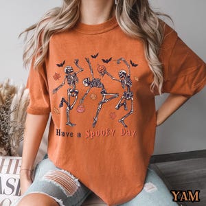 May include: Orange t-shirt featuring a graphic of three skeletons dancing and holding pumpkins. The text "Have a Spooky Day" is printed below the skeletons.