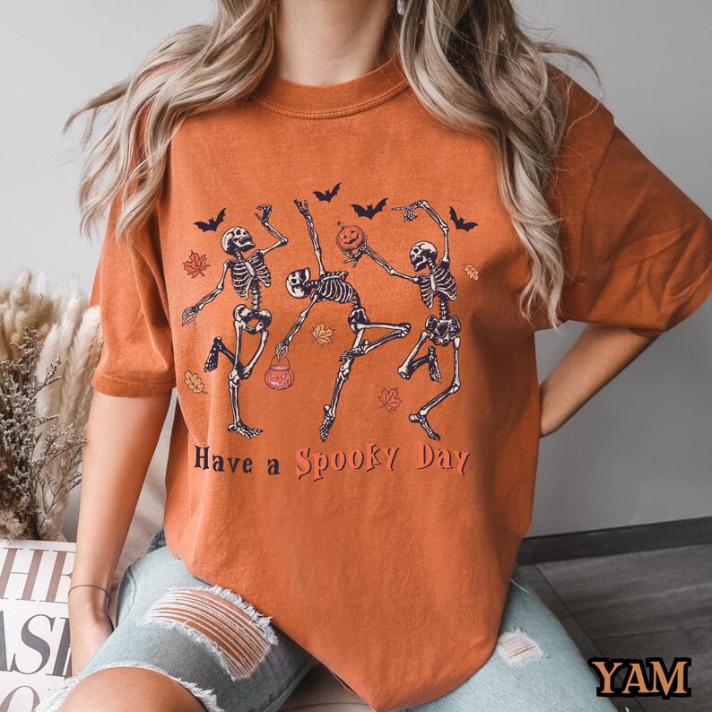 May include: Orange t-shirt featuring a graphic of three skeletons dancing and holding pumpkins. The text "Have a Spooky Day" is printed below the skeletons.