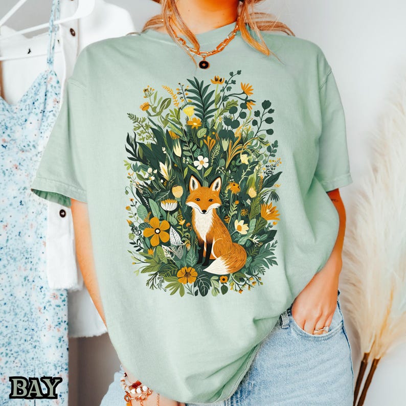May include: A light green t-shirt with a graphic of a fox surrounded by flowers and greenery. The fox is orange and white with a bushy tail.