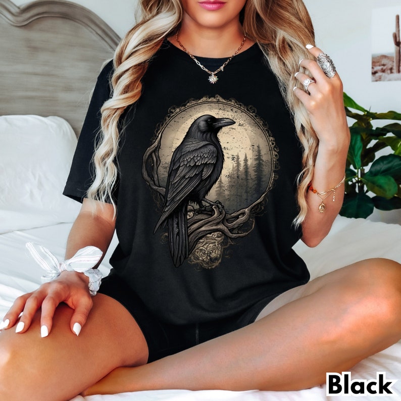May include: Black t-shirt with a graphic of a raven perched on a branch in a moonlit forest. The raven is black with a detailed design and the forest is depicted in shades of gray and brown.