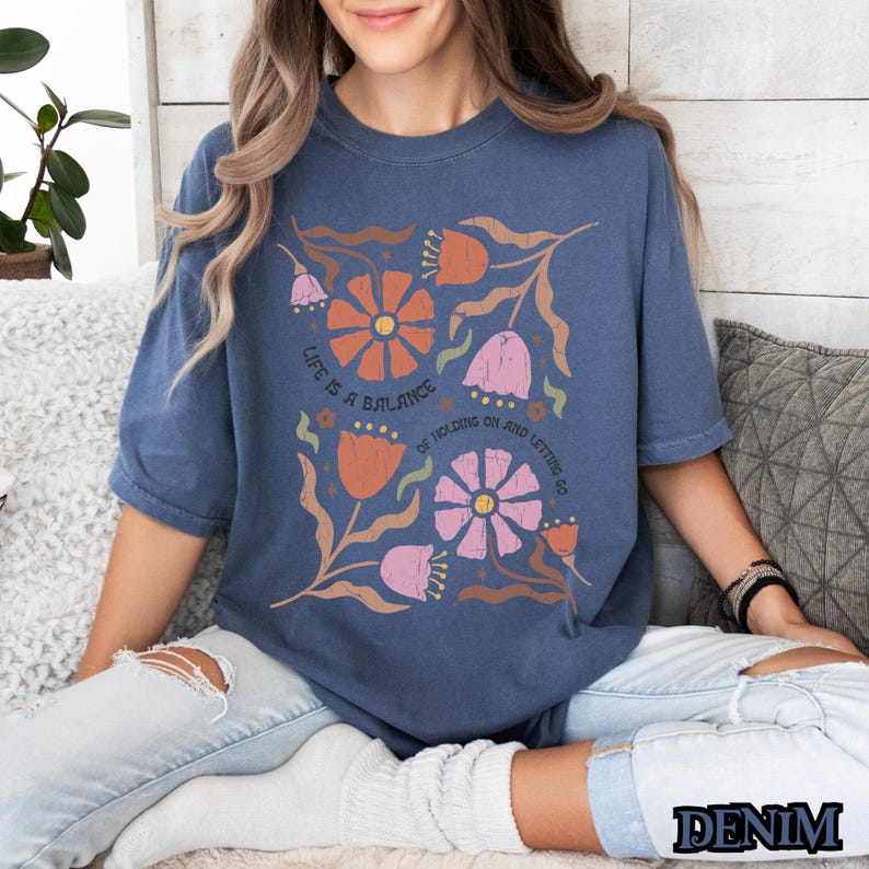 Pu&ograve; includere: Una maglietta blu navy con un design floreale e il testo "Life is a balance of holding on and letting go".