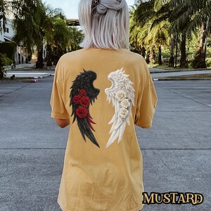May include: A mustard yellow t-shirt with a black and white angel wing design. The wings are on the back of the shirt and are surrounded by red roses.