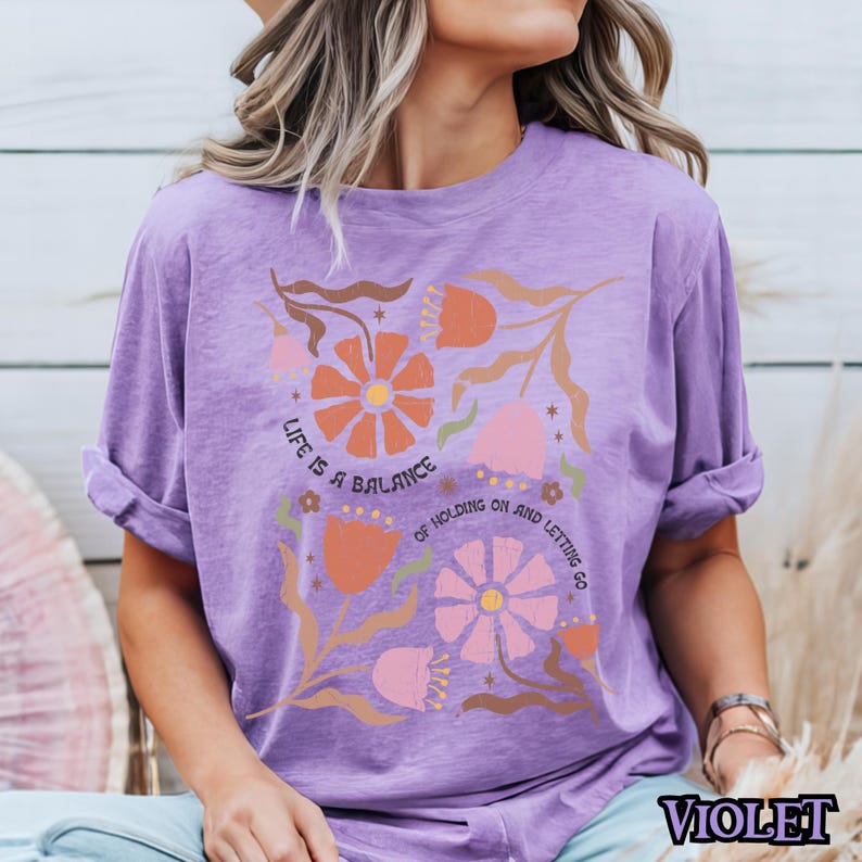 Pu&ograve; includere: Maglietta viola con un design floreale e il testo "Life is a balance of holding on and letting go".
