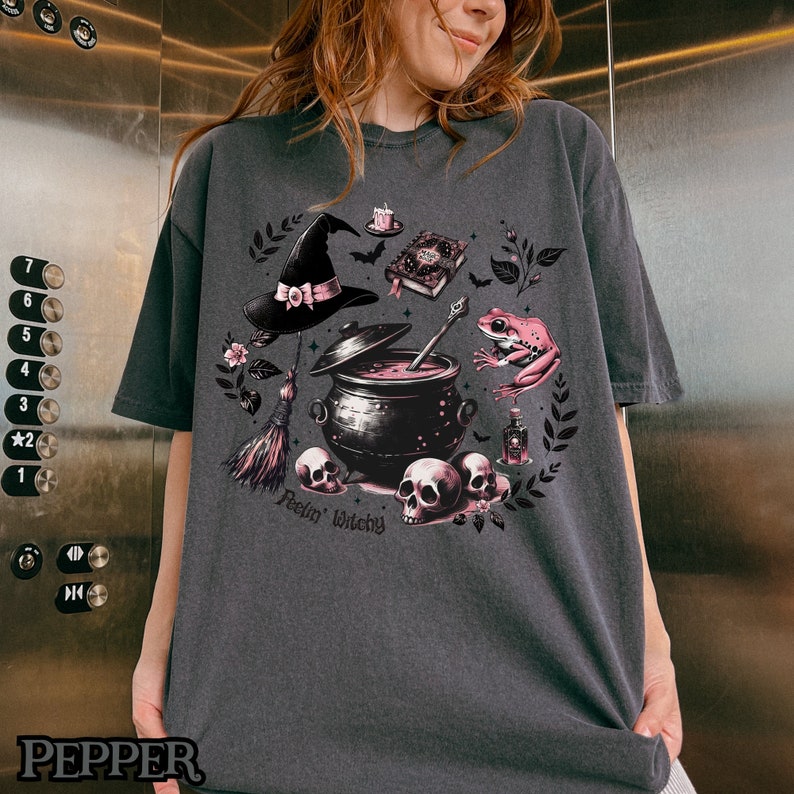 May include: A dark gray t-shirt with a pink and black witchy design. The design features a witch's hat, a cauldron, a frog, a book, a broom, and skulls. The text "Feelin' Witchy" is printed on the shirt.