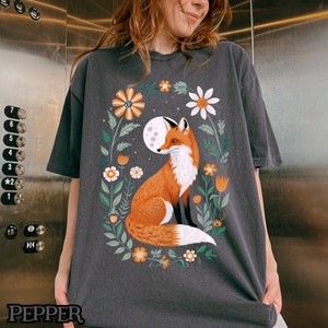 Op de afbeelding: Een donkergrijs T-shirt met een afbeelding van een vos die in een bloemenveld zit. De vos is oranje en wit met een borstelige staart. De bloemen zijn in verschillende kleuren, waaronder roze, geel en blauw.