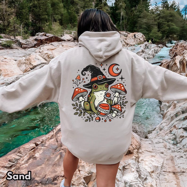 May include: A beige hoodie featuring a graphic print of a frog wearing a witch's hat and holding a potion bottle. The frog is surrounded by mushrooms and stars. The text on the bottle reads "Potion".