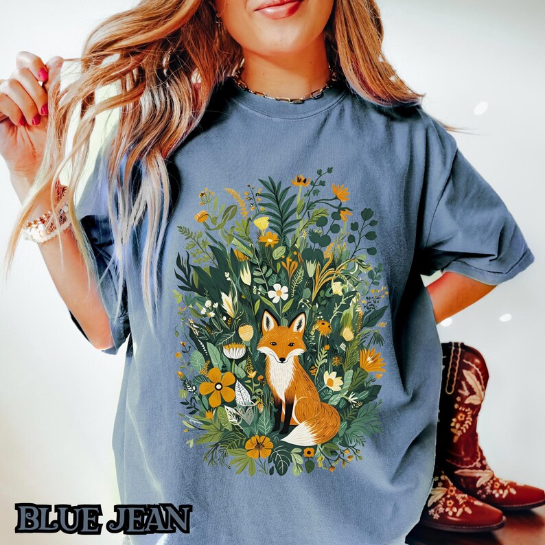 May include: A blue t-shirt with a graphic of a fox surrounded by flowers and greenery. The fox is orange and white with a bushy tail.