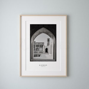 May include: Black and white photograph of a courtyard in Kashan, Iran. The courtyard is framed by a series of arches, and a tall minaret can be seen in the distance.
