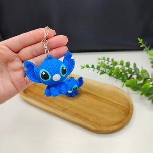 Peut inclure: Un porte-clés Stitch bleu avec un anneau argenté. Le porte-clés présente un design texturé détaillé du personnage avec de grands yeux noirs et blancs. Une plus petite figurine Stitch est également présente. Les articles sont sur un plateau en bois.