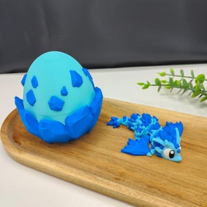 May include: A blue and turquoise dragon hatching from a matching egg. The egg is in a blue nest. The dragon has blue and turquoise scales and wings. The objects are on a wooden tray.