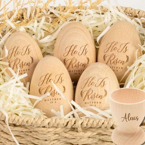 May include: Five wooden Easter eggs in a basket with straw. Each egg is engraved with the words "He Is Risen" and a verse from Matthew. There is also a wooden egg cup with the name "Alexis" engraved on it.