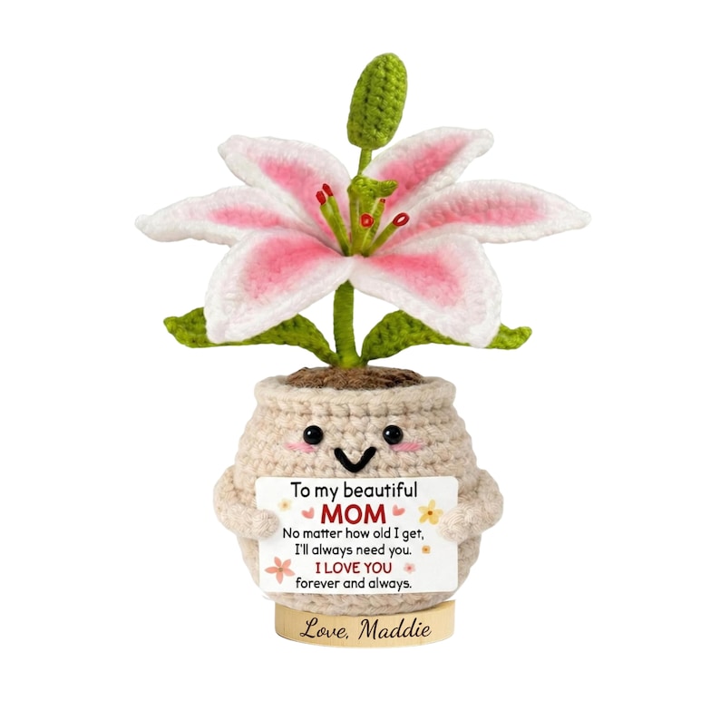 May include: A crocheted lily flower with pink and white petals sits in a smiling pot. The pot holds a sign that reads "To my beautiful MOM" with a loving message and the name "Love, Maddie" at the bottom. The flower is on a wooden base.