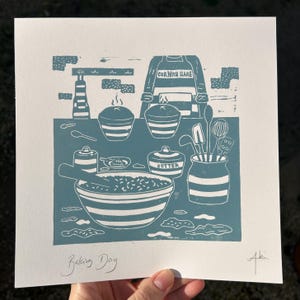 May include: A teal and white linocut print titled "Baking Day" featuring kitchen tools and ingredients. The print includes a bowl, utensils, and the text "CORNISH HARE" on an apron. The print is on white paper.