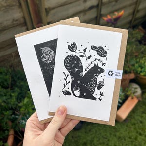 May include: Two greeting cards with a black and white squirrel illustration, held in a hand. The squirrel is surrounded by floral and acorn designs. One card features a black and white seashell design. The cards are in brown envelopes.