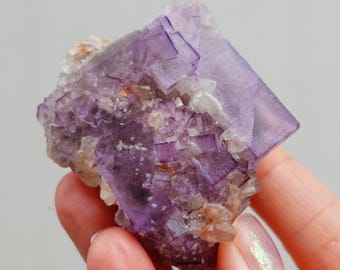 Puede incluir: Un cúmulo de cristales de fluorita púrpura con forma cúbica. Los cristales son translúcidos y tienen un ligero brillo. La fluorita es un mineral natural y tiene una textura rugosa.