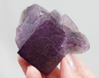 Puede incluir: Un gran cristal de fluorita púrpura con forma cúbica. El cristal tiene una superficie lisa y pulida y es translúcido.