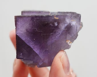 Puede incluir: Un cristal de fluorita púrpura con una forma geométrica y un aspecto en capas. El cristal tiene un color púrpura intenso con algunas vetas de color púrpura más claro y blanco.