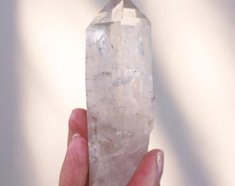 May include: A clear quartz crystal point with a double termination. The crystal is approximately 10 cm tall and has a natural, raw appearance.