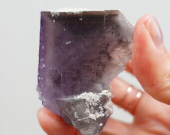 Puede incluir: Primer plano de un cristal de fluorita morado y gris. El cristal tiene forma de cubo con una superficie rugosa y texturizada. Se ven depósitos minerales blancos en los bordes. El cristal se sostiene en una mano.