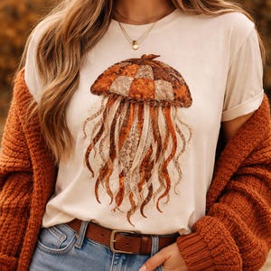 Pode incluir: Camiseta creme com uma ilustração detalhada de uma água-viva. O design da água-viva apresenta um topo xadrez marrom e laranja com tentáculos fluidos em forma de fita em tons de marrom e creme. A camisa é combinada com jeans azuis e um cinto marrom.