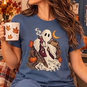 May include: A blue short-sleeve t-shirt featuring a graphic of a white ghost dressed as a medical professional with a stethoscope, clipboard, and coffee cup, surrounded by autumn leaves, a plaid blanket, a pumpkin, and a crescent moon.