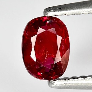 Puede incluir: Una gema de rubí facetada, de forma ovalada, sujeta por unas pinzas plateadas. El rubí es de un color rojo intenso y saturado con reflejos internos. La gema está tallada para maximizar el retorno de la luz.