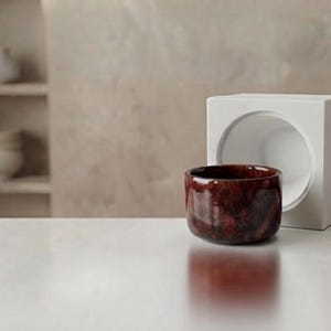 Puede incluir: Un cuenco de cerámica pequeño, redondo y de color marrón rojizo oscuro con un acabado brillante y moteado, situado frente a un objeto blanco en forma de cubo con un gran recorte circular. El cuenco tiene un borde liso y curvo.